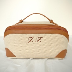 Puede incluir: Un neceser beige con ribetes de cuero marrón y un asa a juego. La bolsa tiene cierre de cremallera y las iniciales bordadas "J.F." en la parte delantera. Las esquinas están reforzadas con cuero marrón.