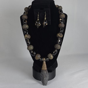 May include: A necklace and earring set displayed on a black jewellery stand. The necklace features large, round, brown and clear beads, with a pendant. The earrings have a similar design. The jewellery is set against a white background.