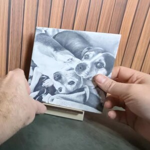 Puede incluir: Una impresión en blanco y negro de dos perros, sostenida frente a un fondo de madera. Los perros están acostados, uno de ellos mirando directamente al espectador. La impresión es cuadrada y parece una fotografía o una impresión sobre una baldosa.