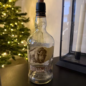 Op de afbeelding: Een heldere glazen Buffalo Trace whiskyfles met een bruine houten stop. De fles heeft een etiket met een afbeelding van een bizon en de woorden "Buffalo Trace". De fles is versierd met lichtslingers.