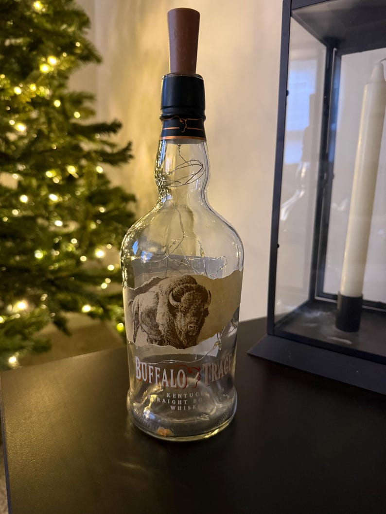Op de afbeelding: Een heldere glazen Buffalo Trace whiskyfles met een bruine houten stop. De fles heeft een etiket met een afbeelding van een bizon en de woorden "Buffalo Trace". De fles is versierd met lichtslingers.