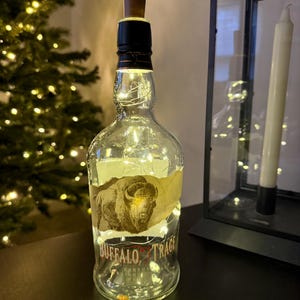 Op de afbeelding: Een hergebruikte Buffalo Trace fles met een houten stop, omwikkeld met lichtsnoeren. De fles heeft een etiket met een afbeelding van een bizon en de merknaam. De lichten cre&euml;ren een warme, decoratieve lichtbron.