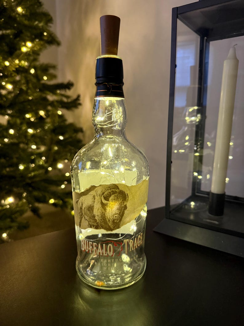 Op de afbeelding: Een hergebruikte Buffalo Trace fles met een houten stop, omwikkeld met lichtsnoeren. De fles heeft een etiket met een afbeelding van een bizon en de merknaam. De lichten cre&euml;ren een warme, decoratieve lichtbron.