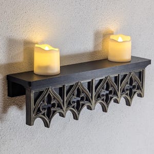 May include: A black wooden shelf with a gothic-style design. Two battery-operated candles with a warm glow sit on the shelf. The shelf is mounted on a textured, off-white wall.