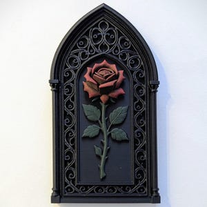 Op de afbeelding: Een zwart gothic-stijl wandkunstwerk met een gedetailleerd reliëf van een roos. De roos is dieprood met groene bladeren en stengel, tegen een zwarte achtergrond in een boogvormig frame.
