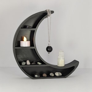 May include: A black crescent moon shelf with multiple compartments. It holds a small candle, various stones, a pendulum, and a small glass bottle. The shelf is designed for displaying crystals and spiritual items.