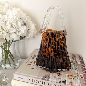 May include: A glass vase shaped like a handbag with a clear handle and a brown and black leopard print pattern. The vase has a fluted, pleated body and sits on top of a book titled A HOME to SHARE by Leslie Saeta, which is stacked on another book.