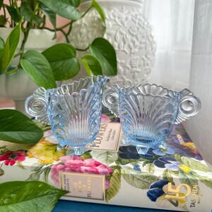 May include: Two light blue glass vessels with a scalloped rim, ribbed vertical detailing, and small feet. Each piece features a circular handle on either side. The glass has a translucent quality and a shell-like pattern throughout the body.