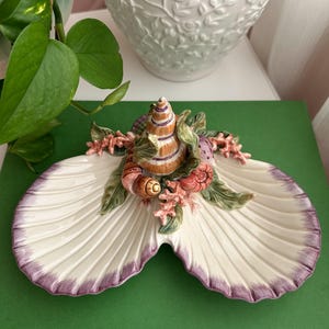 May include: A ceramic seashell-shaped dish with a white interior and a purple-edged rim. The dish is adorned with a central cluster of seashells, coral, and green leaves. The decorative piece is set on a green surface.