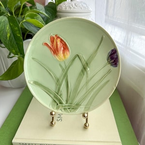 May include: A round, light green ceramic plate featuring a raised, embossed design of a single orange and yellow tulip and a smaller purple tulip with green stems and leaves. The plate is displayed on a small gold-toned metal stand.