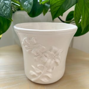 May include: A white ceramic plant pot with a flared rim and a raised, embossed floral design on the front. The surface of the pot has a smooth, glossy finish, and the relief pattern features detailed petals and leaves integrated into the body.