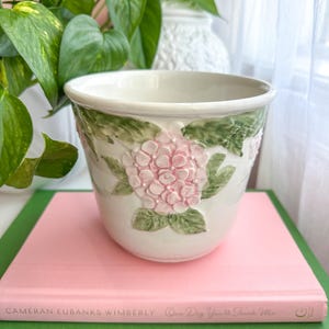 May include: A white ceramic planter featuring a raised, three-dimensional relief of pink hydrangea flowers and green leaves around the exterior. The pot has a smooth, glossy finish and a wide, circular opening with a slightly flared rim.