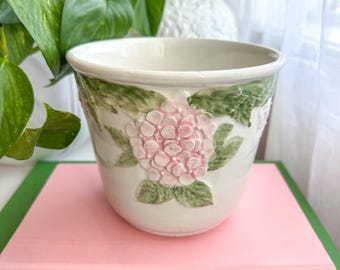 May include: A white ceramic planter featuring a raised, three-dimensional relief of pink hydrangea flowers and green leaves around the exterior. The pot has a smooth, glossy finish and a wide, circular opening with a slightly flared rim.