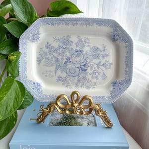 May include: An off-white rectangular serving platter with a blue floral pattern and a decorative border. It's displayed on a gold-coloured metal stand, resting on a light blue book with the word "Joie" on the cover.