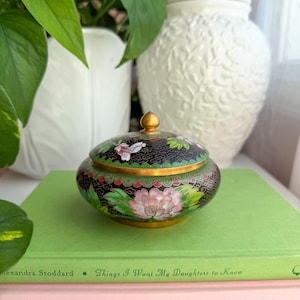 May include: A round cloisonné trinket box with a lid featuring a black background, gold metal trim, and a floral pattern in shades of pink, green, and white. The lid has a small gold finial at the center, and the body displays intricate enamel work.