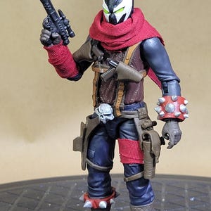 May include: Action figure of a character in a black and white mask with green eyes, wearing a red scarf, brown vest, and blue pants. The figure is holding a black gun and has spiked wrist and ankle bands.
