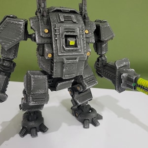 May include: A gray, armored robot figurine with a boxy torso, articulated limbs, and a large weapon attachment on one arm. The figure features metallic paint, small gold-colored circular details, and a bright neon green glowing effect on the weapon.