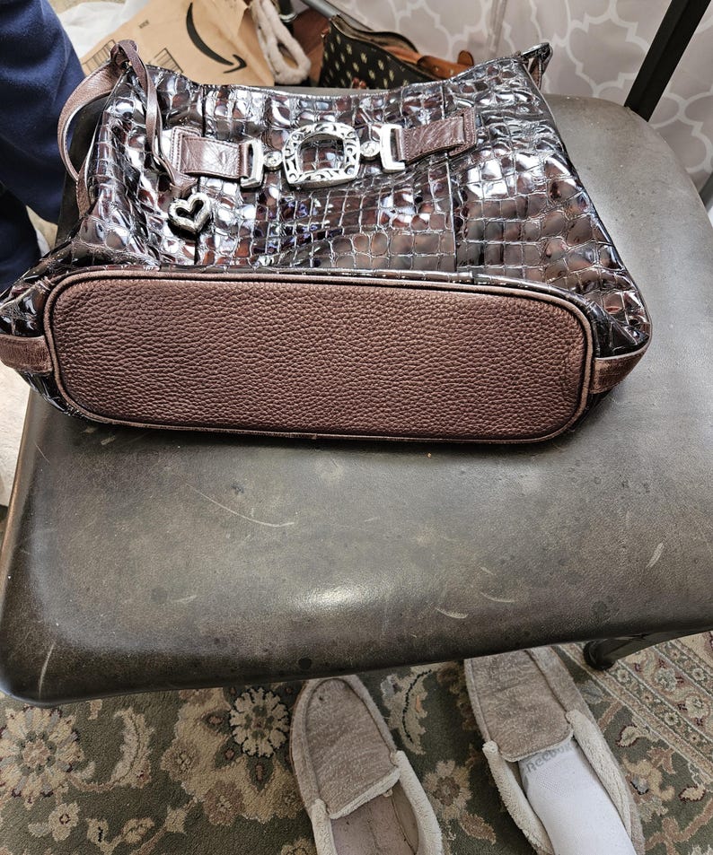 K&ouml;nnte beinhalten: Eine braune, gl&auml;nzende Handtasche mit Krokodilmuster. Die Tasche hat einen strukturierten braunen Boden und dekorative silberne Schnallen. Ein kleiner herzf&ouml;rmiger Anh&auml;nger h&auml;ngt vorne. Die Tasche steht auf einem dunklen Metallstuhl.