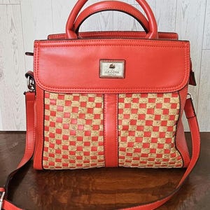 May include: A red handbag with a woven cork design on the front. The bag has a top handle and a detachable shoulder strap. The brand name "J.R. Cork" is displayed on a small metal plate.