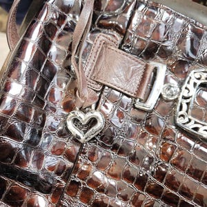 K&ouml;nnte beinhalten: Braune, gl&auml;nzende Handtasche mit Krokodilleder-Muster. Die Tasche hat einen herzf&ouml;rmigen Silberanh&auml;nger, einen Lederriemen und eine silberne Schnalle. Die Tasche ist mittelgro&szlig; und hat ein gl&auml;nzendes Finish.