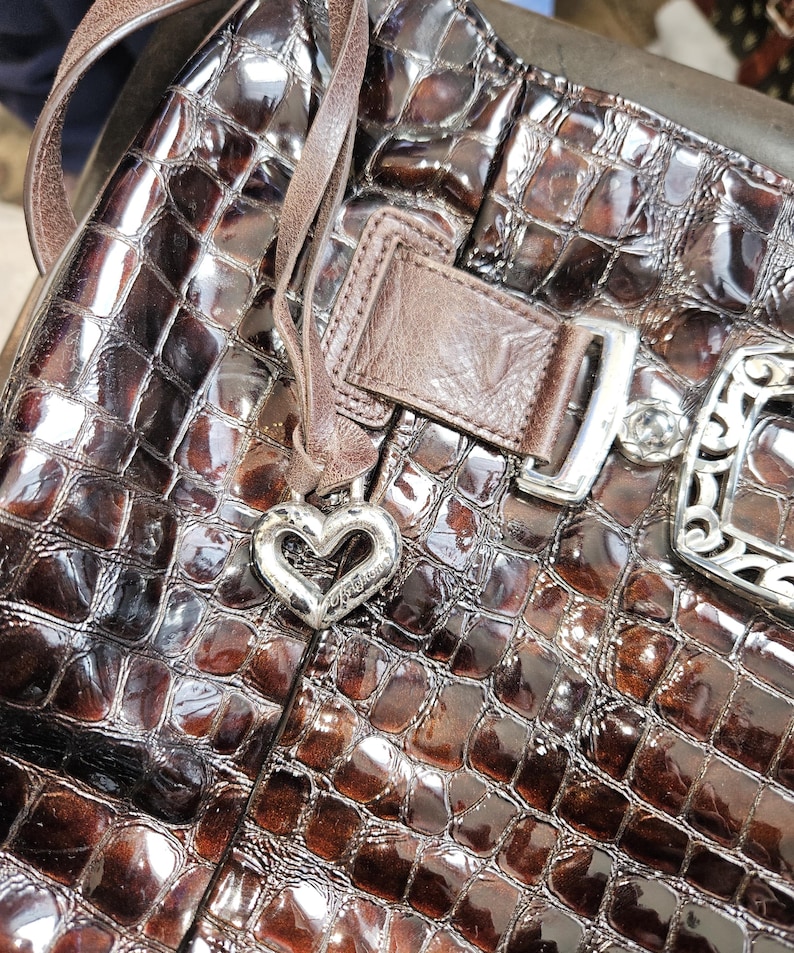 K&ouml;nnte beinhalten: Braune, gl&auml;nzende Handtasche mit Krokodilleder-Muster. Die Tasche hat einen herzf&ouml;rmigen Silberanh&auml;nger, einen Lederriemen und eine silberne Schnalle. Die Tasche ist mittelgro&szlig; und hat ein gl&auml;nzendes Finish.