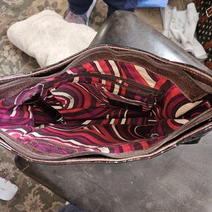 K&ouml;nnte beinhalten: Offene Ansicht einer braunen Lederhandtasche mit farbenfrohem Innenraum. Der Innenraum weist ein lebendiges Muster aus roten, wei&szlig;en und lila gebogenen Linien auf. Die Tasche hat mehrere Taschen und einen Rei&szlig;verschluss.