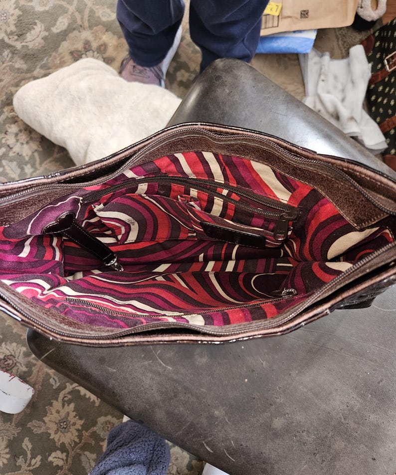 K&ouml;nnte beinhalten: Offene Ansicht einer braunen Lederhandtasche mit farbenfrohem Innenraum. Der Innenraum weist ein lebendiges Muster aus roten, wei&szlig;en und lila gebogenen Linien auf. Die Tasche hat mehrere Taschen und einen Rei&szlig;verschluss.