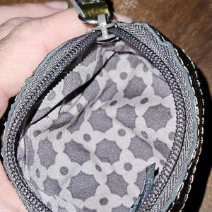 May include: A small, open coin purse with a zipper closure. The exterior is a dark gray with a metallic sheen, and the interior features a gray and white geometric pattern. The zipper pull is a dark metallic color.