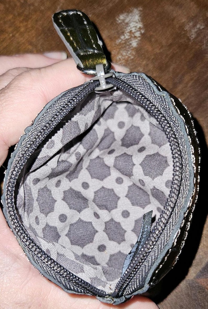 May include: A small, open coin purse with a zipper closure. The exterior is a dark gray with a metallic sheen, and the interior features a gray and white geometric pattern. The zipper pull is a dark metallic color.