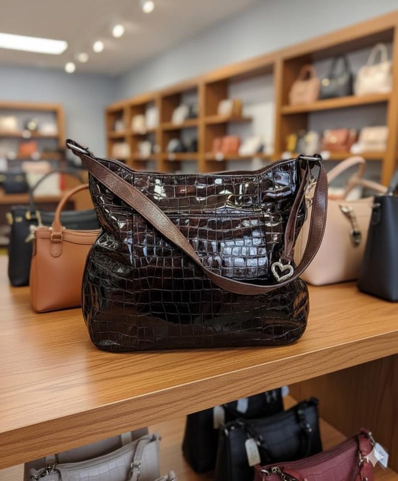 K&ouml;nnte beinhalten: Eine gl&auml;nzende, dunkelbraune Handtasche mit Krokodilhautstruktur liegt auf einem Holzregal. Die Tasche hat einen braunen Lederriemen und einen herzf&ouml;rmigen Silberanh&auml;nger. Weitere Handtaschen sind im Hintergrund zu sehen.