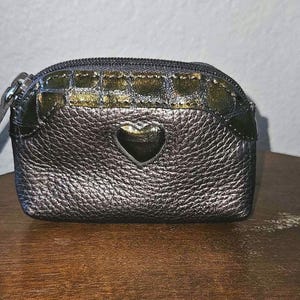 May include: A small, metallic silver coin purse with a heart-shaped cutout. The purse has a textured surface and a gold and green patterned top section. It features a zipper closure and a small leather pull.