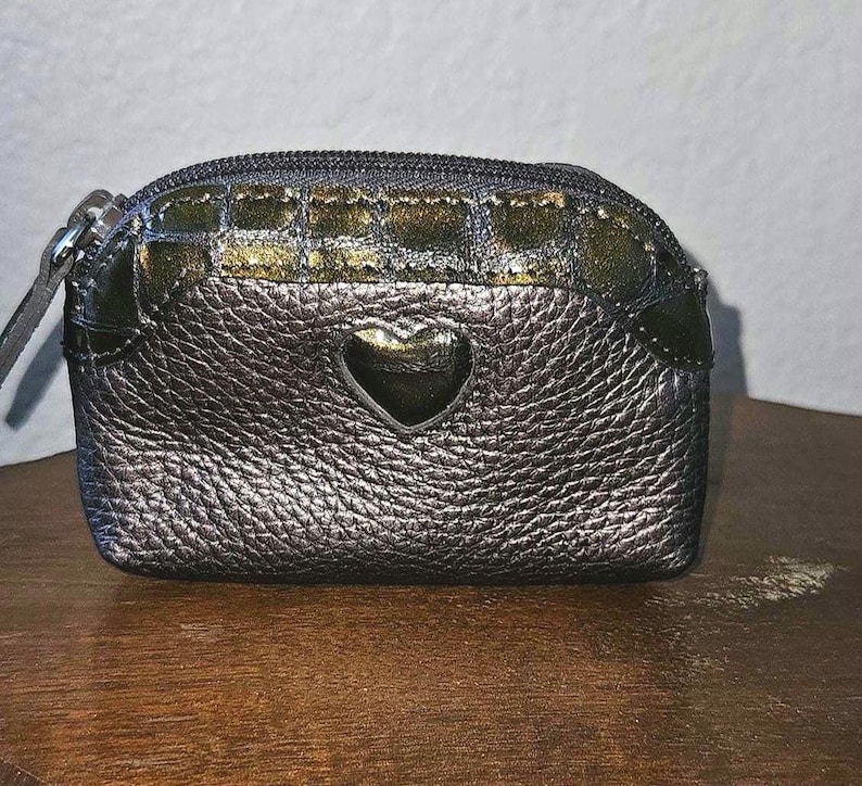 May include: A small, metallic silver coin purse with a heart-shaped cutout. The purse has a textured surface and a gold and green patterned top section. It features a zipper closure and a small leather pull.