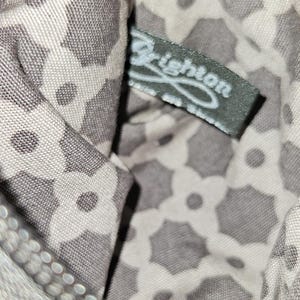 May include: Close-up of a small, patterned pouch with a zipper. The interior fabric features a repeating floral design in gray and white. A tag with the brand name "Brighton" is visible inside the pouch.