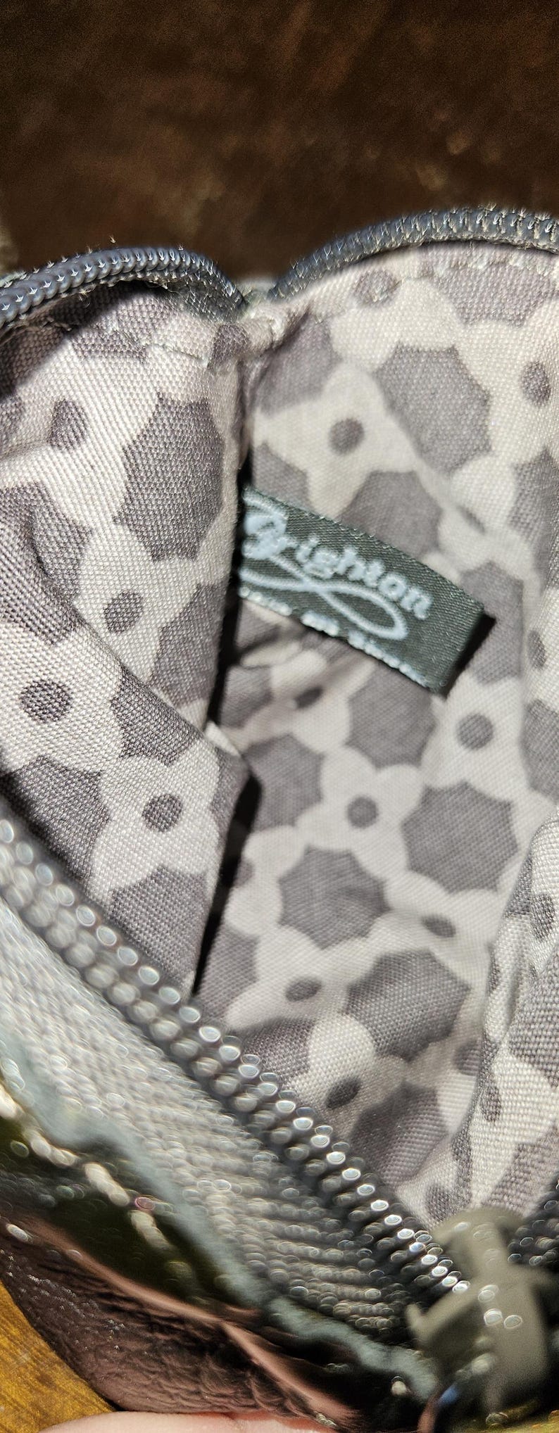 May include: Close-up of a small, patterned pouch with a zipper. The interior fabric features a repeating floral design in gray and white. A tag with the brand name "Brighton" is visible inside the pouch.