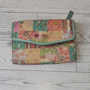 May include: A patchwork wallet with a flap closure. The wallet features a variety of floral and geometric patterns in shades of pink, green, and yellow. The edges are trimmed with a light green material and studded with small silver rivets. A small green tab is attached.