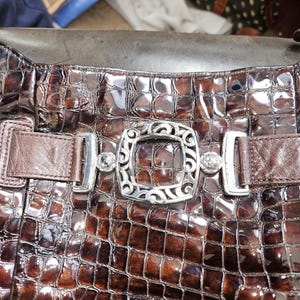 K&ouml;nnte beinhalten: Nahaufnahme einer braunen, strukturierten Handtasche mit Krokodilmuster. Die Tasche hat eine dekorative silberne Schnalle mit aufwendigen Details und zwei silbernen Verschl&uuml;ssen. Die Tasche hat braune Lederriemen.