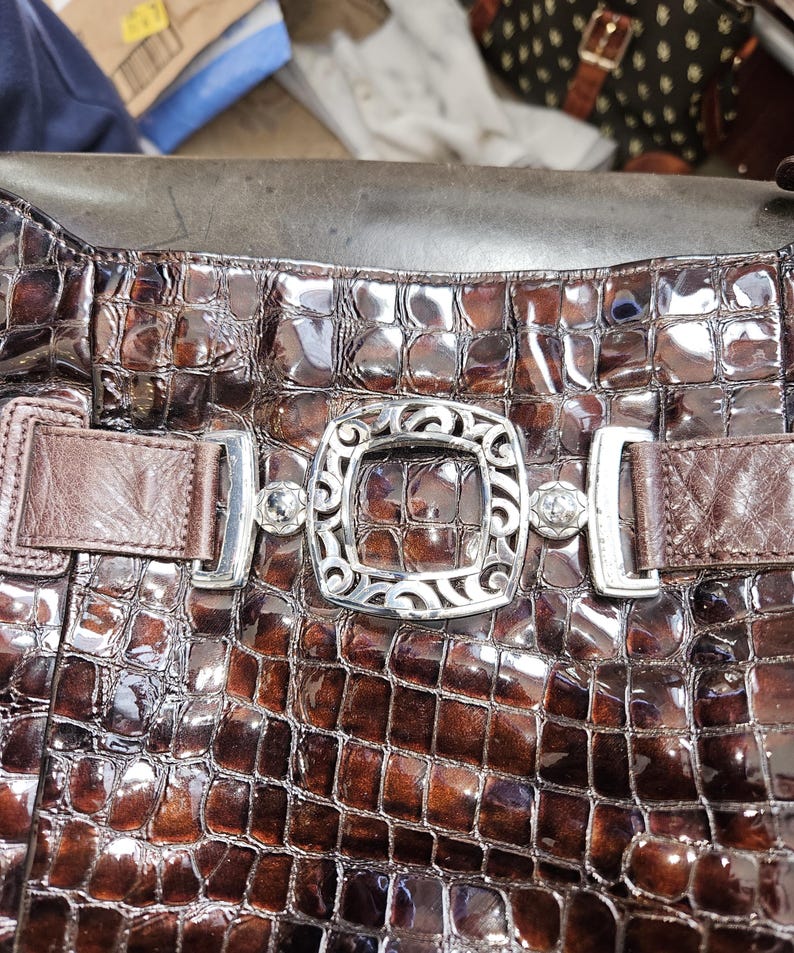 K&ouml;nnte beinhalten: Nahaufnahme einer braunen, strukturierten Handtasche mit Krokodilmuster. Die Tasche hat eine dekorative silberne Schnalle mit aufwendigen Details und zwei silbernen Verschl&uuml;ssen. Die Tasche hat braune Lederriemen.