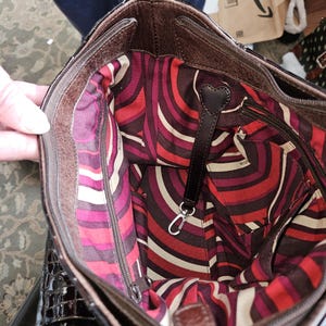 K&ouml;nnte beinhalten: Eine braune Lederhandtasche mit einem gemusterten Innenraum in Rot-, Wei&szlig;- und Lilat&ouml;nen. Die Tasche hat einen Rei&szlig;verschluss und einen schwarzen Lederriemen mit herzf&ouml;rmigem Detail. Das Innenfutter hat ein einzigartiges, abstraktes Design.