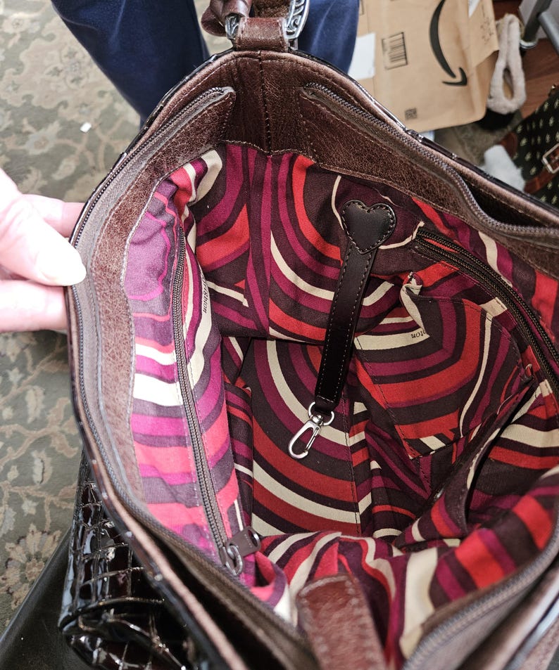 K&ouml;nnte beinhalten: Eine braune Lederhandtasche mit einem gemusterten Innenraum in Rot-, Wei&szlig;- und Lilat&ouml;nen. Die Tasche hat einen Rei&szlig;verschluss und einen schwarzen Lederriemen mit herzf&ouml;rmigem Detail. Das Innenfutter hat ein einzigartiges, abstraktes Design.