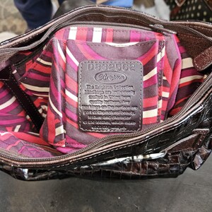 K&ouml;nnte beinhalten: Braune Brighton Handtasche mit gl&auml;nzendem, strukturiertem &Auml;u&szlig;eren. Das Innere hat ein farbenfrohes, gemustertes Futter in Rosa-, Rot- und Wei&szlig;t&ouml;nen. Ein Lederetikett im Inneren zeigt den Markennamen und Produktdetails.