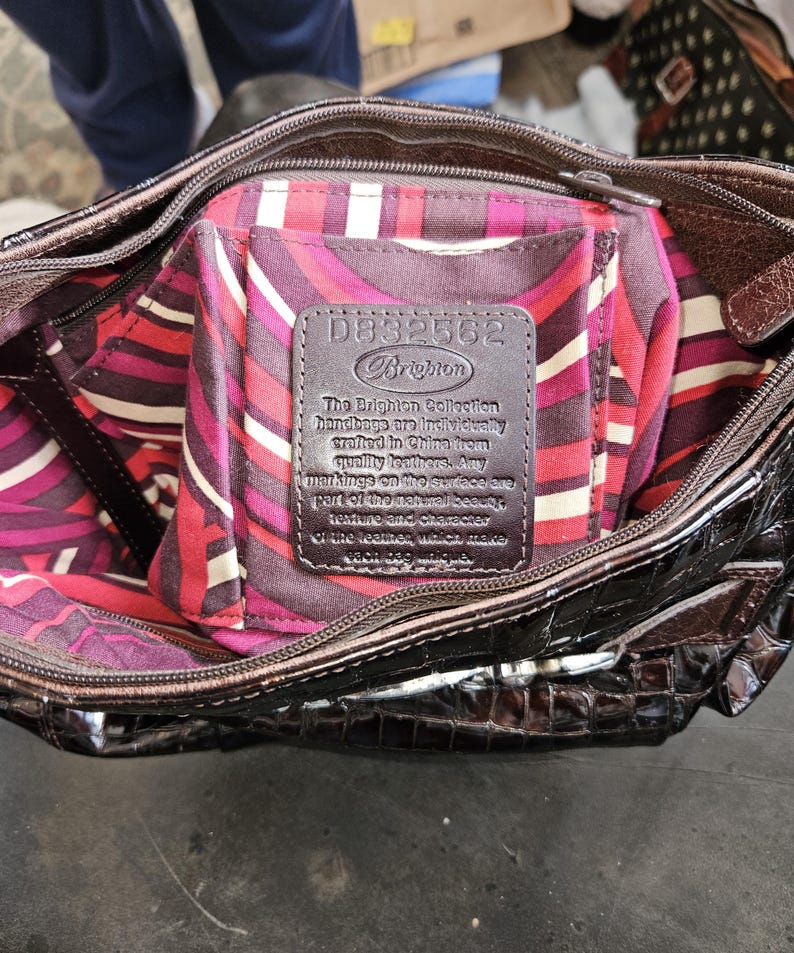 K&ouml;nnte beinhalten: Braune Brighton Handtasche mit gl&auml;nzendem, strukturiertem &Auml;u&szlig;eren. Das Innere hat ein farbenfrohes, gemustertes Futter in Rosa-, Rot- und Wei&szlig;t&ouml;nen. Ein Lederetikett im Inneren zeigt den Markennamen und Produktdetails.