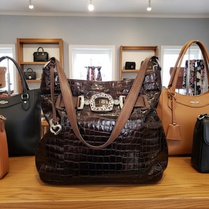 K&ouml;nnte beinhalten: Eine dunkelbraune, strukturierte Lederhandtasche mit silberner Schnalle und braunen Riemen. Die Tasche hat einen herzf&ouml;rmigen Anh&auml;nger und wird auf einem Holzregal mit anderen Handtaschen ausgestellt.