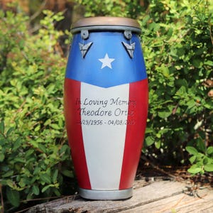 May include: A memorial urn shaped like a conga drum, painted in the colors of the Puerto Rican flag: blue, white, and red. The urn has a bronze-colored top and base. Text on the urn reads "In Loving Memory Theodore Ortiz 12/29/1956 - 04/08/2020".
