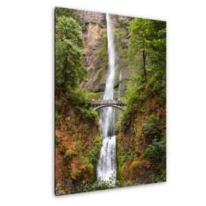 May include: A scenic photograph of a waterfall cascading down a rocky cliff, surrounded by lush green trees. A small arched bridge spans the waterfall, with people visible on it. The image captures the natural beauty of the landscape.