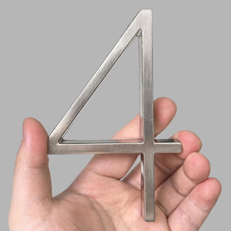 May include: A silver metal number four held in a hand. The number is a stylized font with a triangular top and a straight vertical line. The metal has a brushed finish and is set against a gray background.