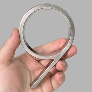 May include: A brushed metal number nine house address plaque. The numeral is held in a hand against a gray background. The number is approximately 5 inches tall.