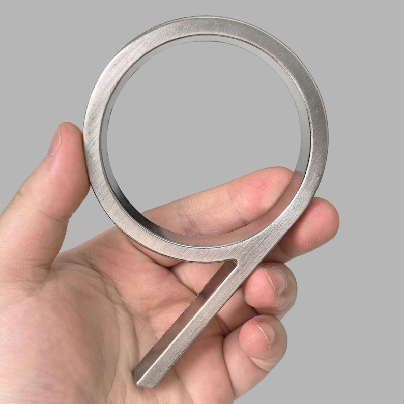May include: A brushed metal number nine house address plaque. The numeral is held in a hand against a gray background. The number is approximately 5 inches tall.