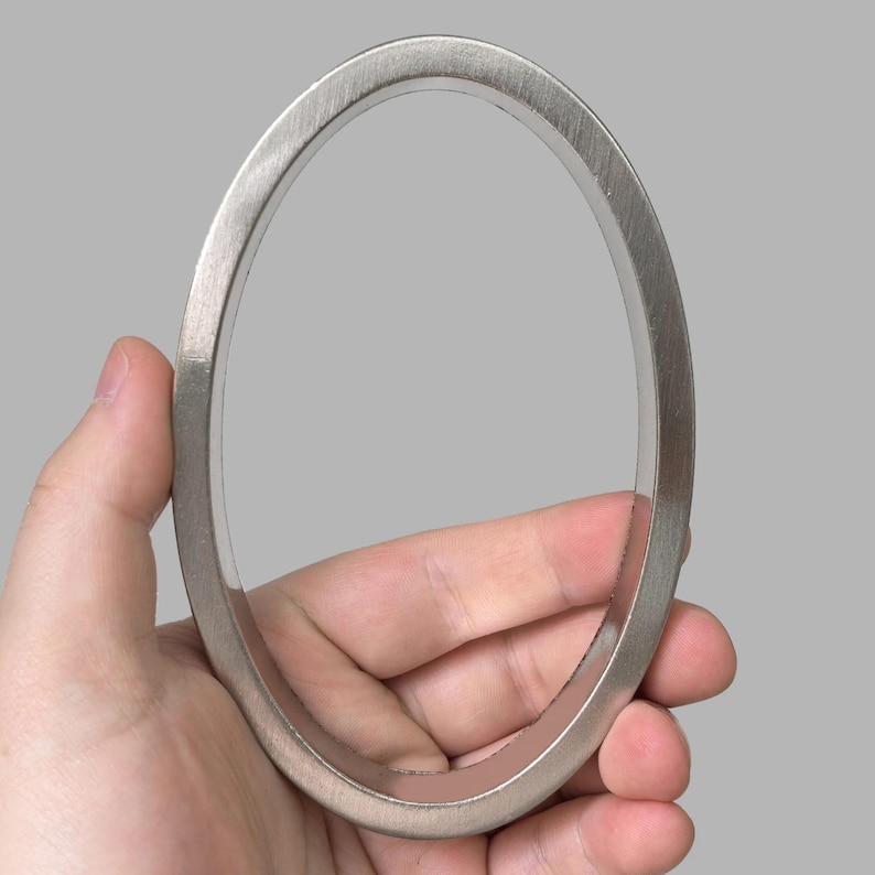 May include: A brushed metal oval-shaped ring held in a hand. The ring has a smooth, matte finish and a simple, minimalist design. The ring is likely a decorative element or part of a larger piece.