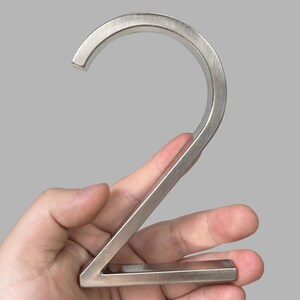 May include: A brushed metal number two, held in a hand. The number has a modern, minimalist design with a curved top and a straight diagonal line. The metal has a matte finish and a silver color.