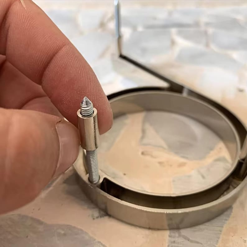 May include: Close-up of a silver-toned metal picture frame with a screw and a pointed tip. The frame has a circular design with a smaller inner circle. The screw is held near the frame, suggesting assembly or repair.